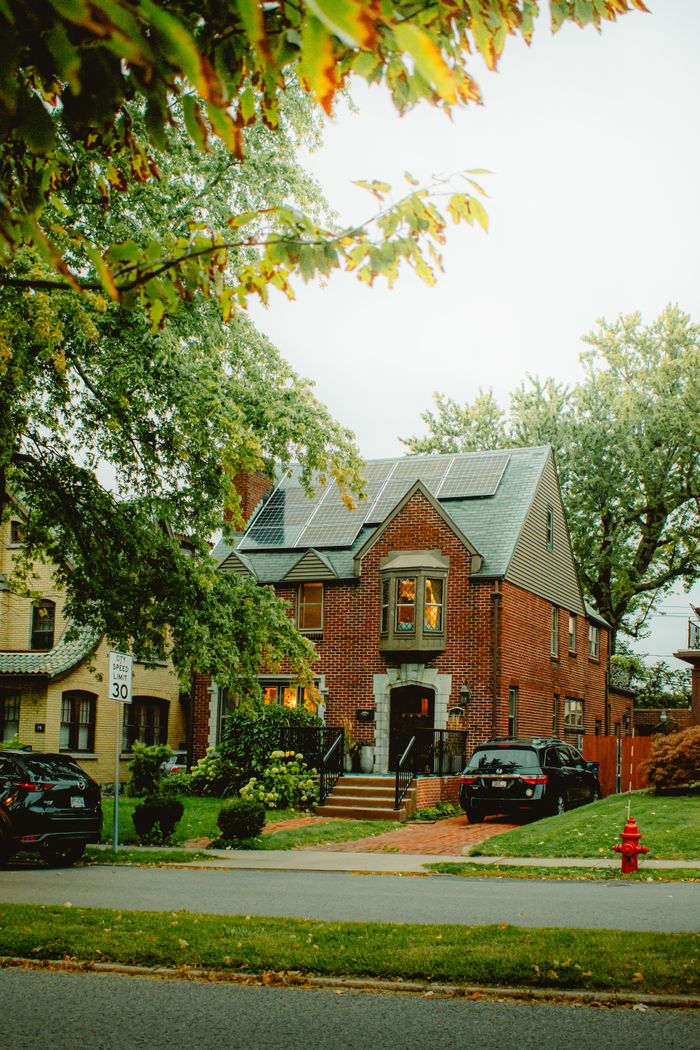hero A picturesque brick house with solar panels, lush greenery, and fall foliage in a suburban neighborhood.